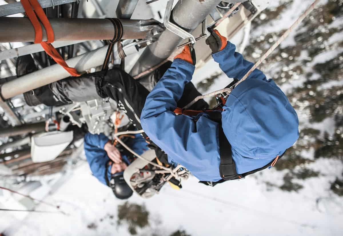 Two people in blue jacket and harnesses working on communication lines.