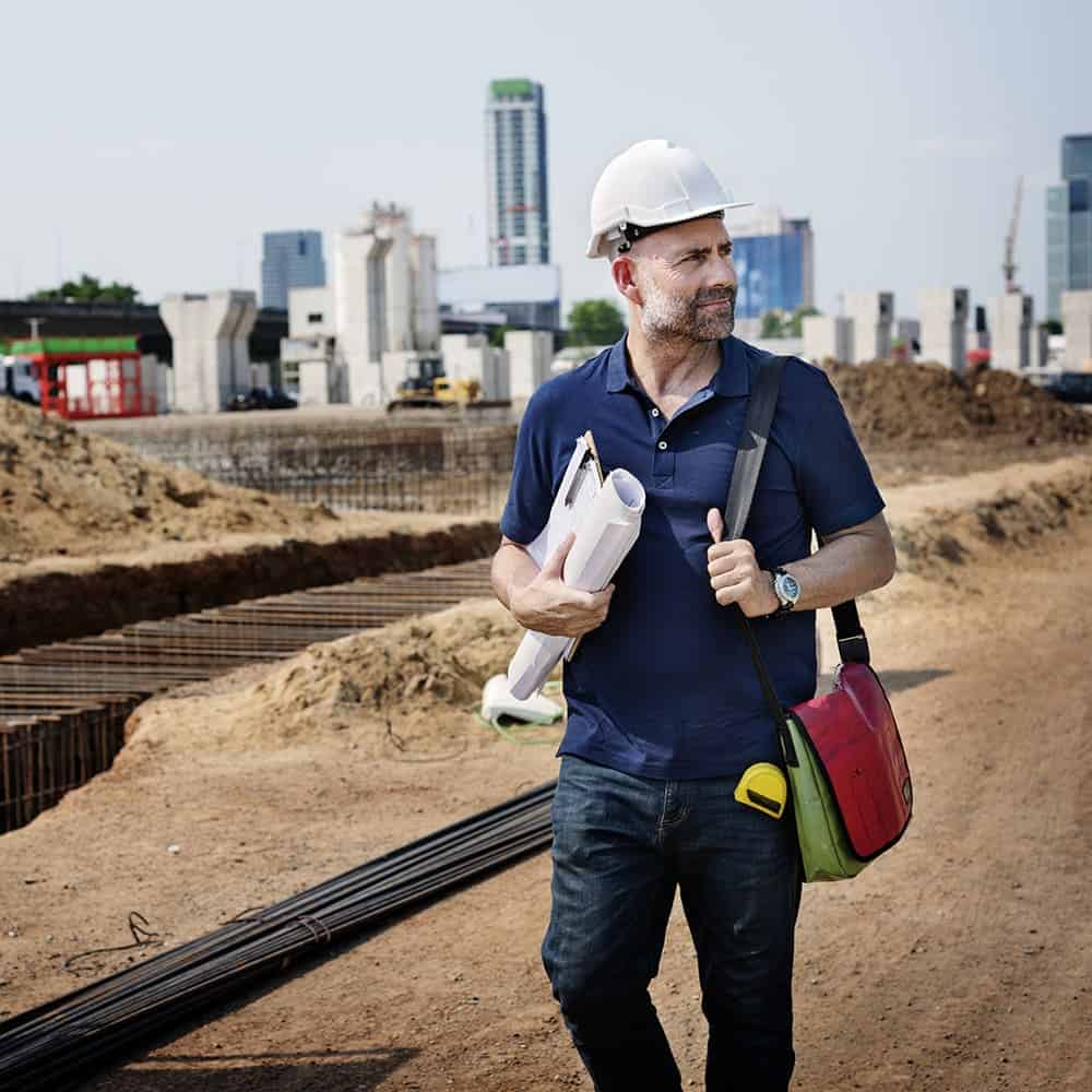 A happy male architect on a dirt covered construction site with blueprints in hand, a bag over his shoulder, a white hard cap on his head, and the city skyline in the background.