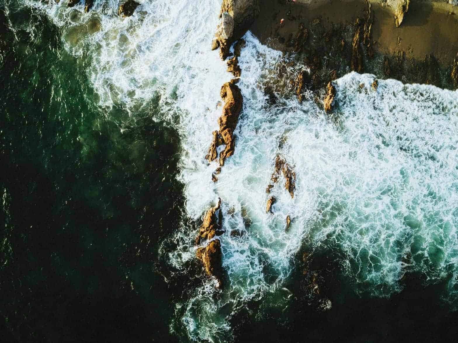 A bird's eye view of a rocky ocean with whitewashed waves and blue water.