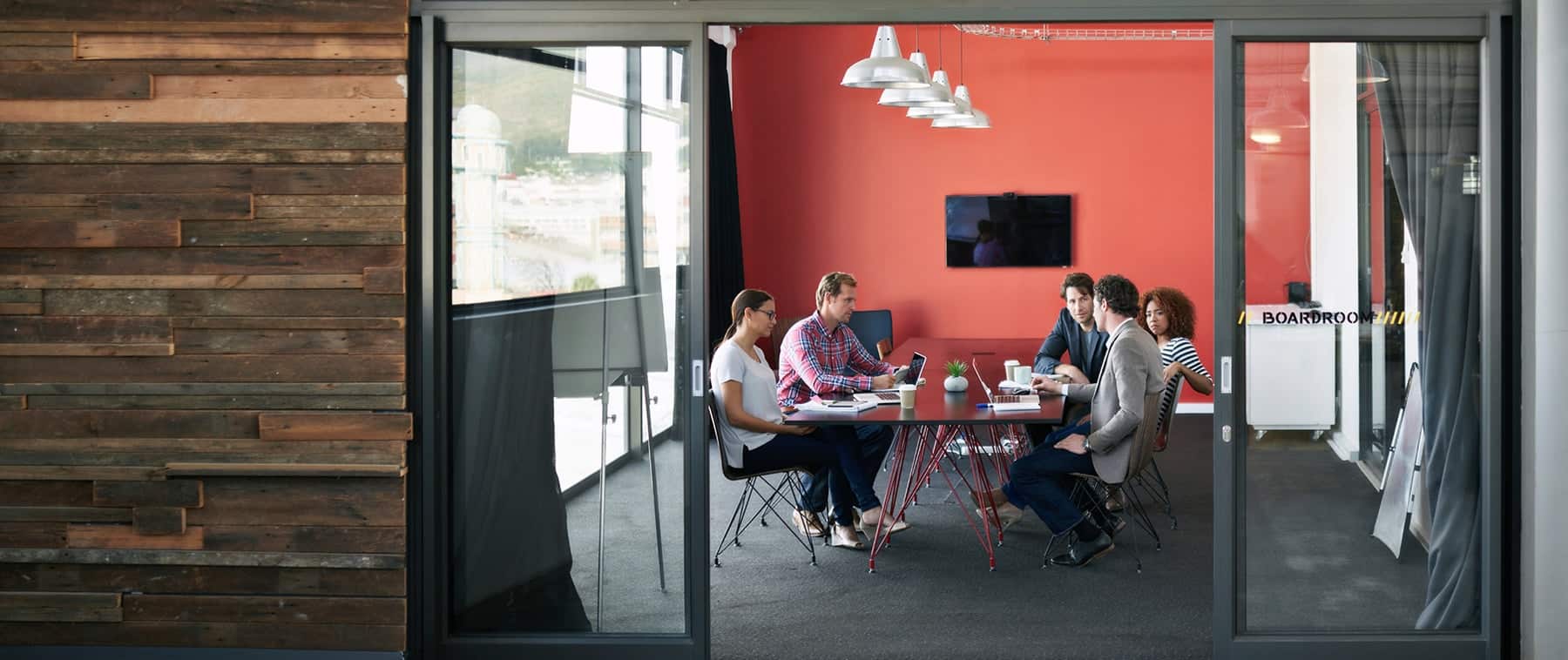 A group of colleagues collaborating around a meeting table in a coral-colored office conference room.