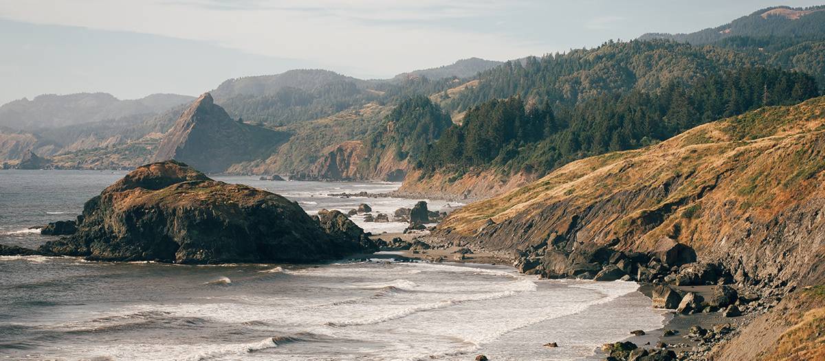 A mountainous coast with a foggy image of the rocky shoreline on the left, with green mountains in the background.