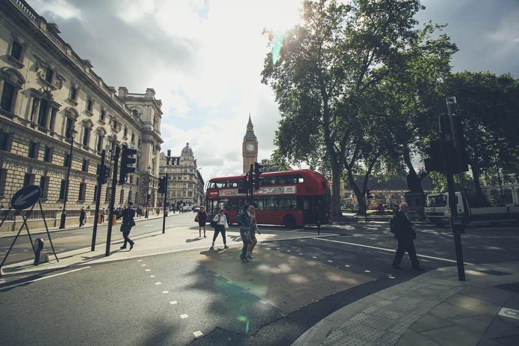 A photo in London with people walking by, a red double decker bus, and with Big Ben in the background during a sunny day.