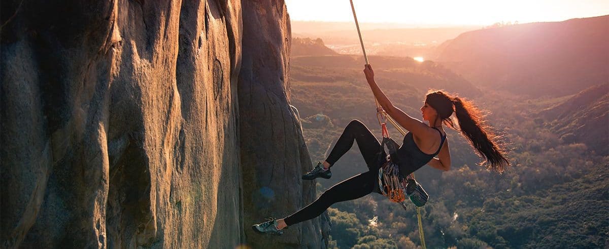 A woman attached to a rope while climbing a cliff with the landscape in the background at sunset.