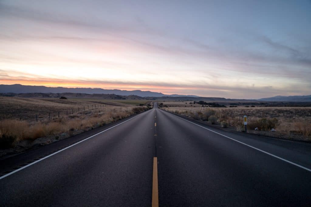 The middle of a road in a grassy area with the mountains and the sunrise in the background.