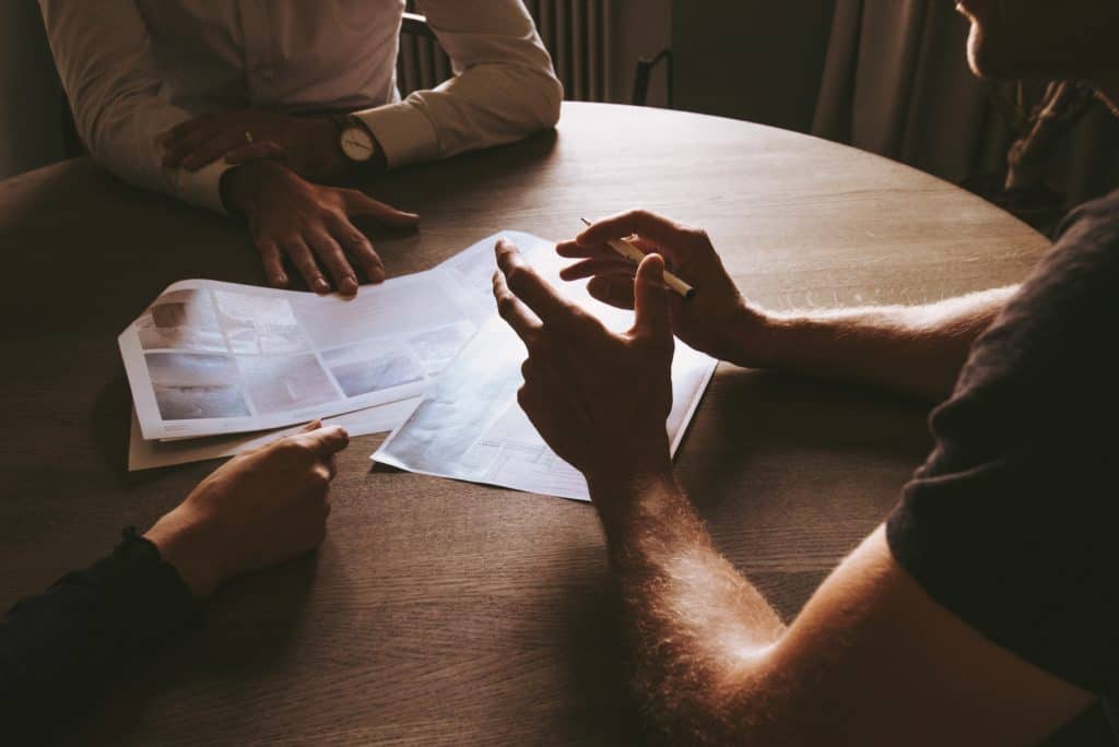 Two people discussing a plan with papers in the middle of a round table in a dark office space.