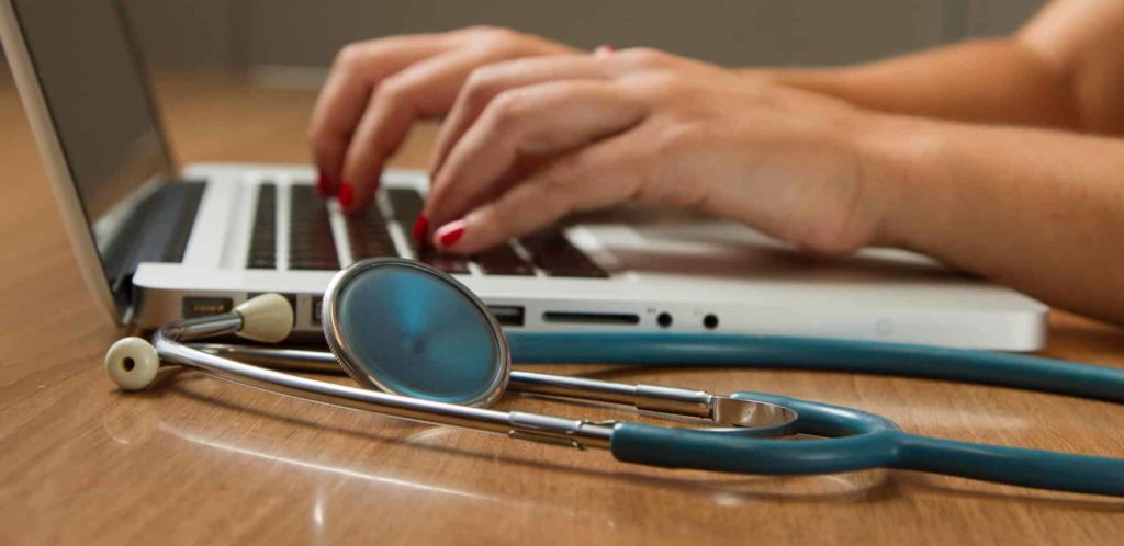 A woman typing on a laptop with a blue stethoscope. Employers with medical plans need to be aware of the newly released IRS inflation-adjusted amounts for 2022 relating to Heath Saving Accounts (HSAs) and high deductible health plans (HDHPs).