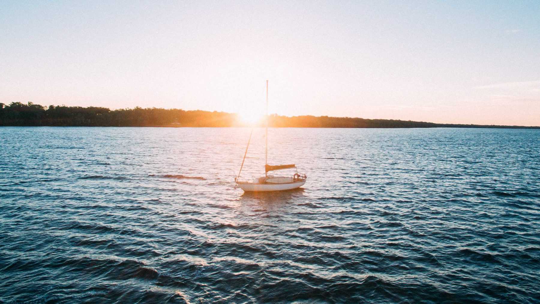 A white sailboat on the water with a strip of land in the background and the sun setting from behind the sail.