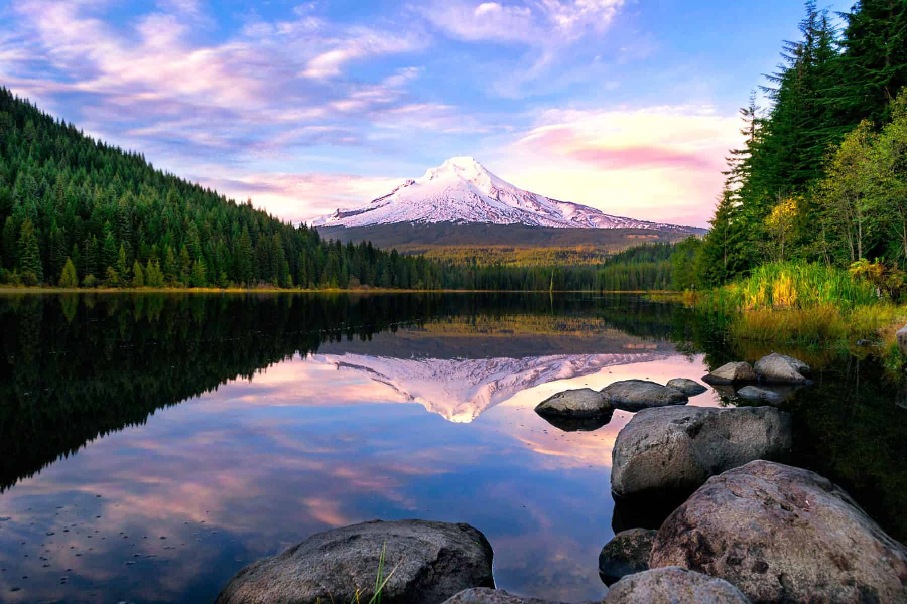 A view of a river and Mount Hood in the distance, with rocks and evergreen trees surrounding the still-reflective river.