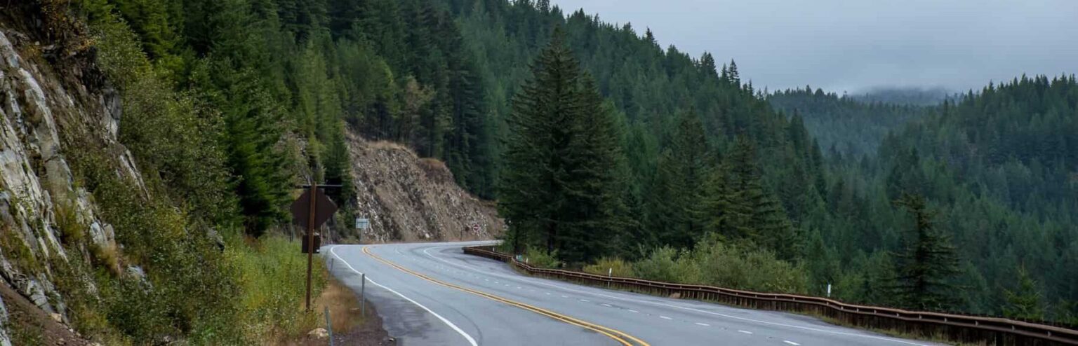 A landscape of a windy road in the foggy mountains surrounded by mountains and green evergreen trees.