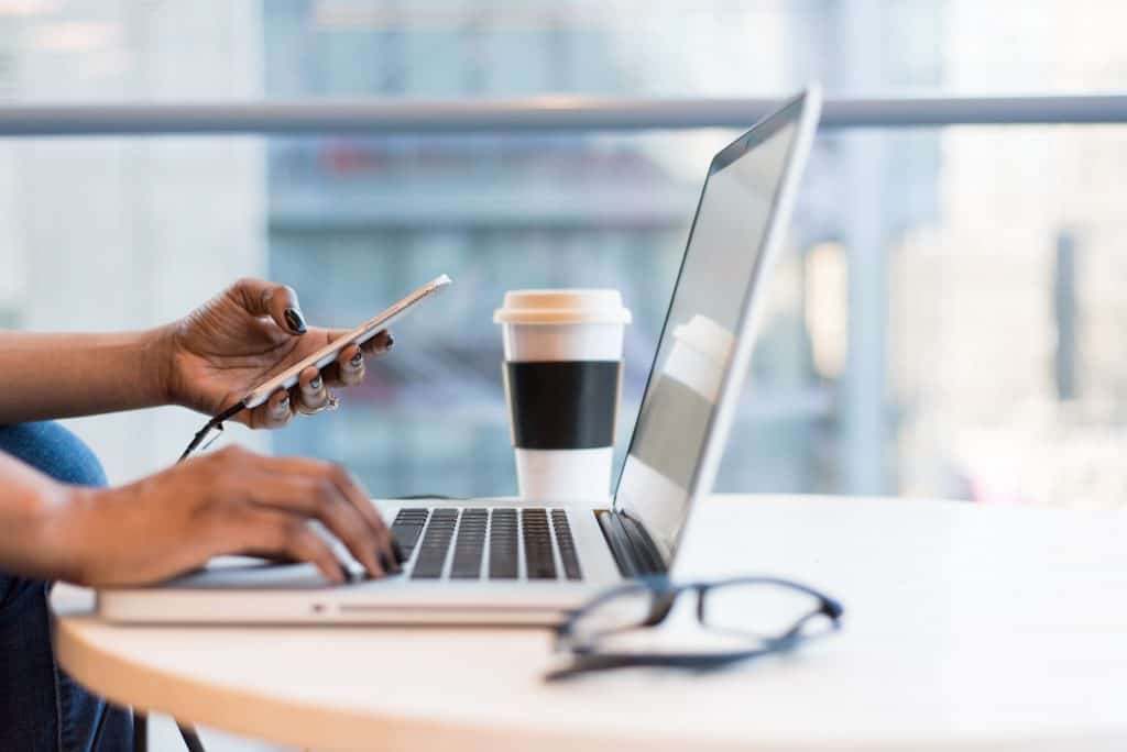 A woman holding her phone while typing on her laptop with a coffee to her left in a brightly lit office space. Business owners everywhere are seeing a record number of mergers and acquisitions (M&A) completed in the first quarter of 2021.