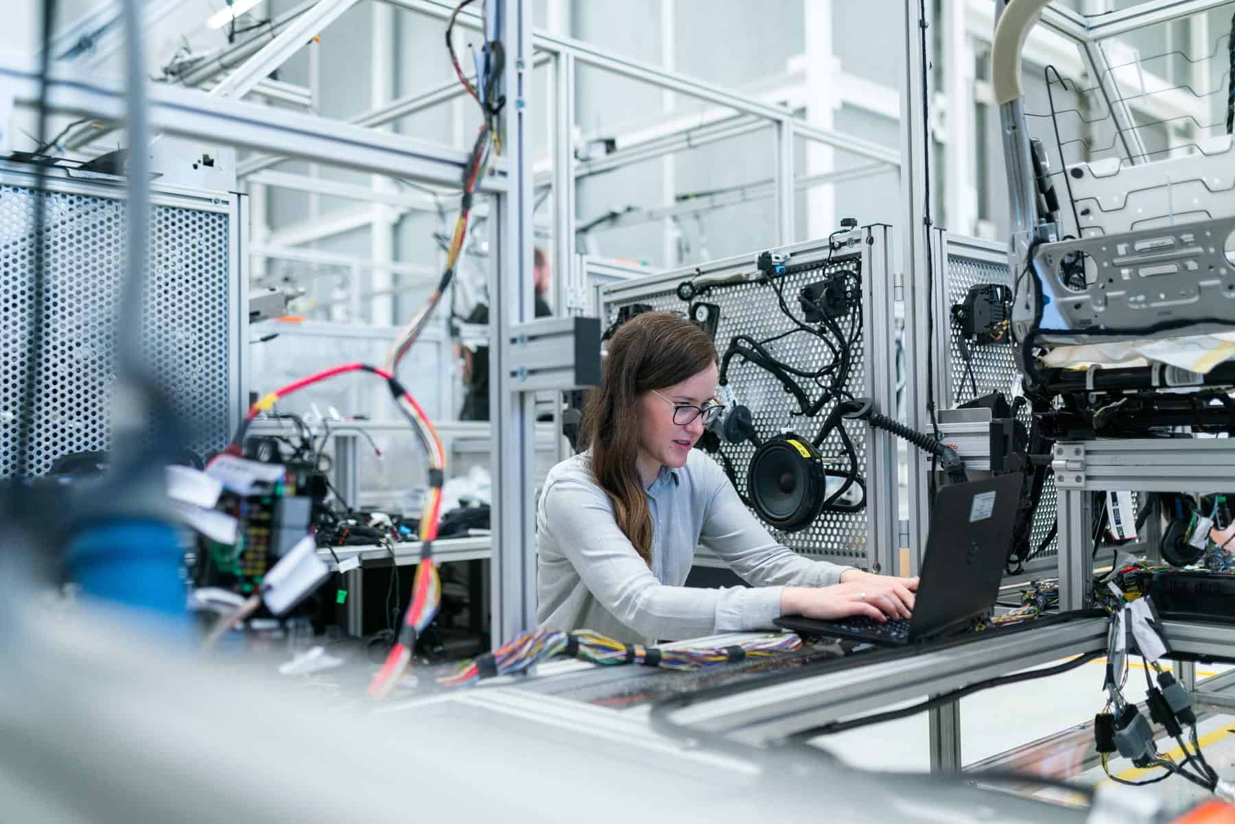 Woman in white long sleeve shirt works on targeted technology application on a black laptop in a manufacturing plant