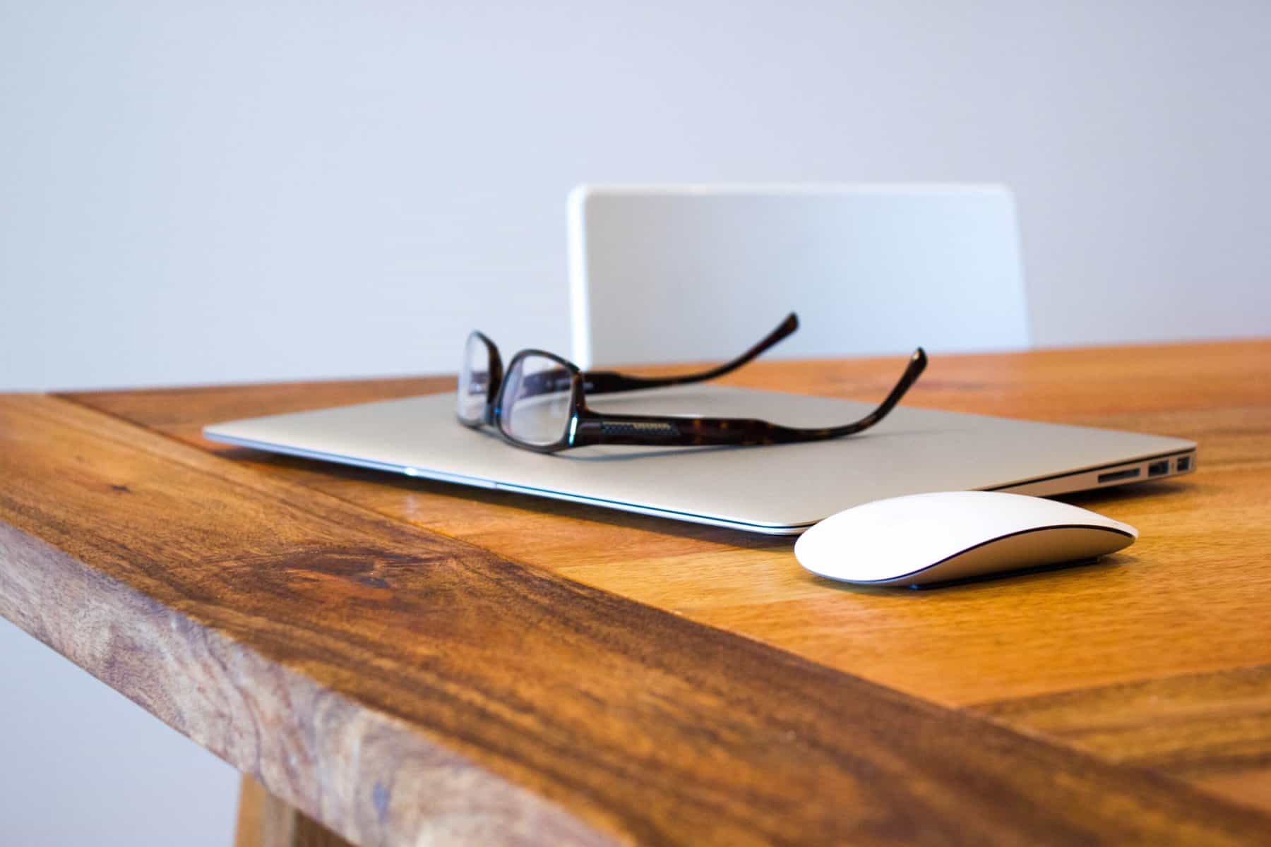 Eyeglasses on top of a closed laptop next to a computer mouse on a wooden table with a white chair in the background