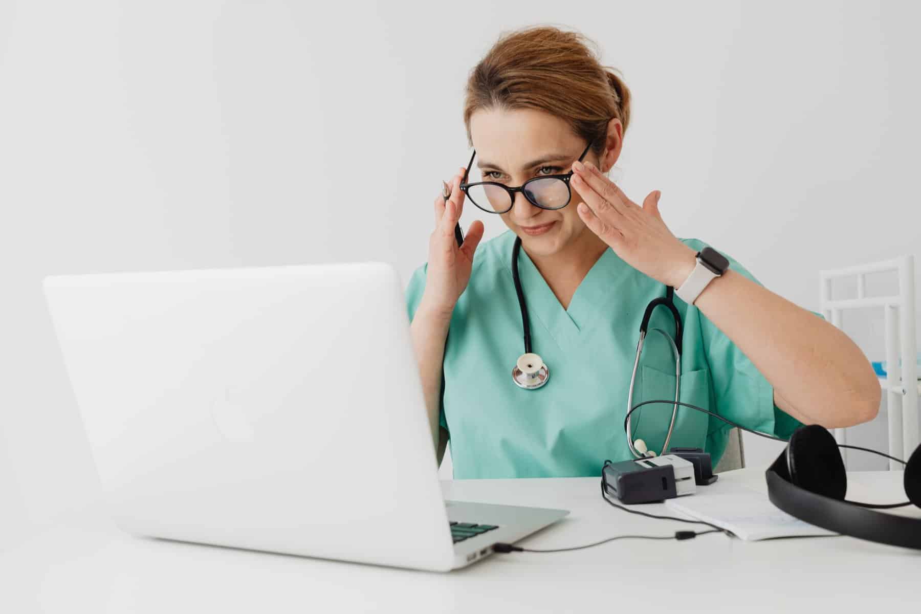 woman in green scrubs with a stethoscope sits in front of white computer