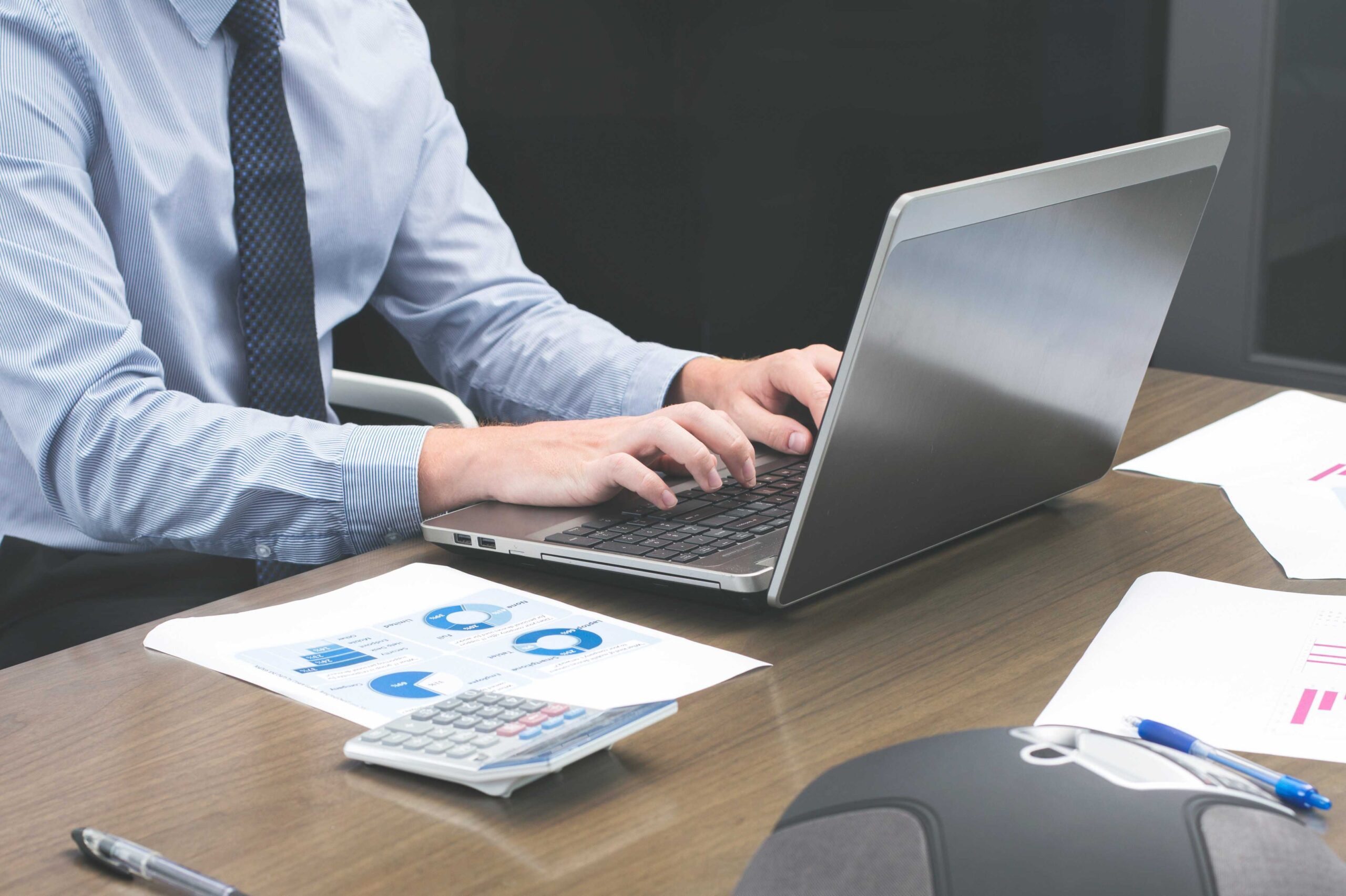 business man sits at desk with his hands on a silver laptop and market forecasts are printed and on the desk